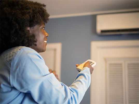 A homeowner using the remote for their heat pump with a heat pump on the wall nearby